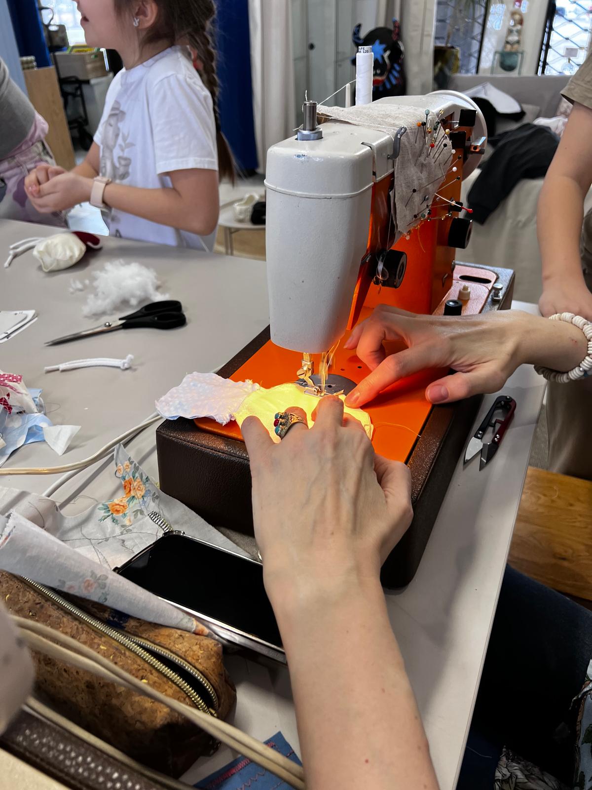 Dziewczyna szyje na maszynie, pracując z żółtą tkaniną na stole pełnym materiałów i narzędzi krawieckich.