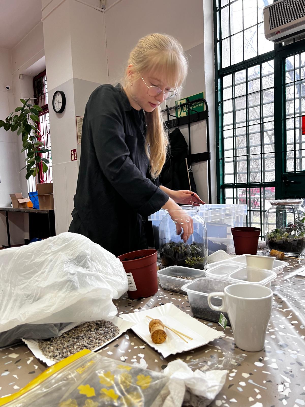 Kobieta w czarnej bluzie pracuje przy stole z roślinami i materiałami w jasnym pomieszczeniu z dużymi oknami.