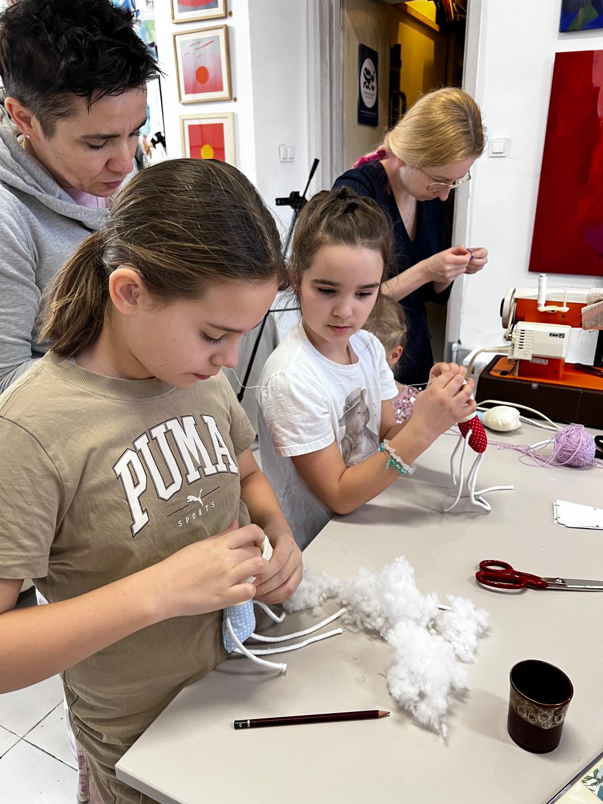 Dzieci uczestniczą w zajęciach rękodzielniczych, tworząc ozdoby z bawełny i kolorowych nici pod opieką dorosłych.