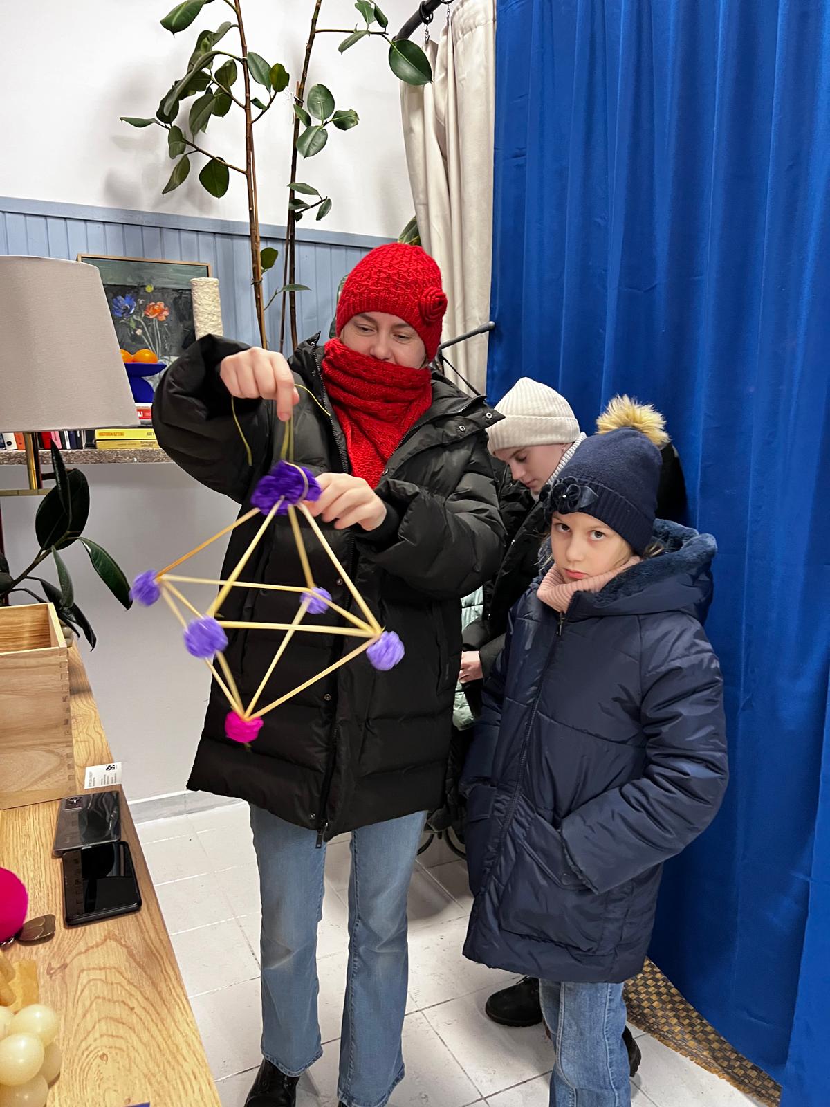 Dorosła osoba w czerwonej czapce pokazuje dzieciom geometryczną strukturę z żółtych patyczków i fioletowych kul.
