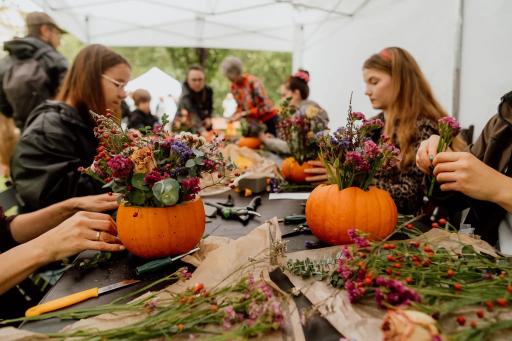 Grupa osób wykonuje jesienne dekoracje kwiatowe w dynach w namiocie podczas kreatywnego warsztatu.
