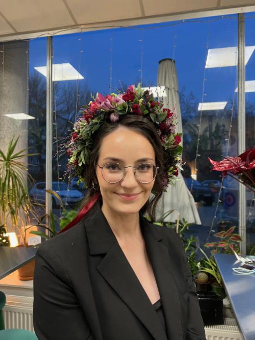 Kobieta w okularach i czarnym żakiecie nosi kwiatową koronę i pozuje w nowoczesnym biurze z błękitnym oknem.