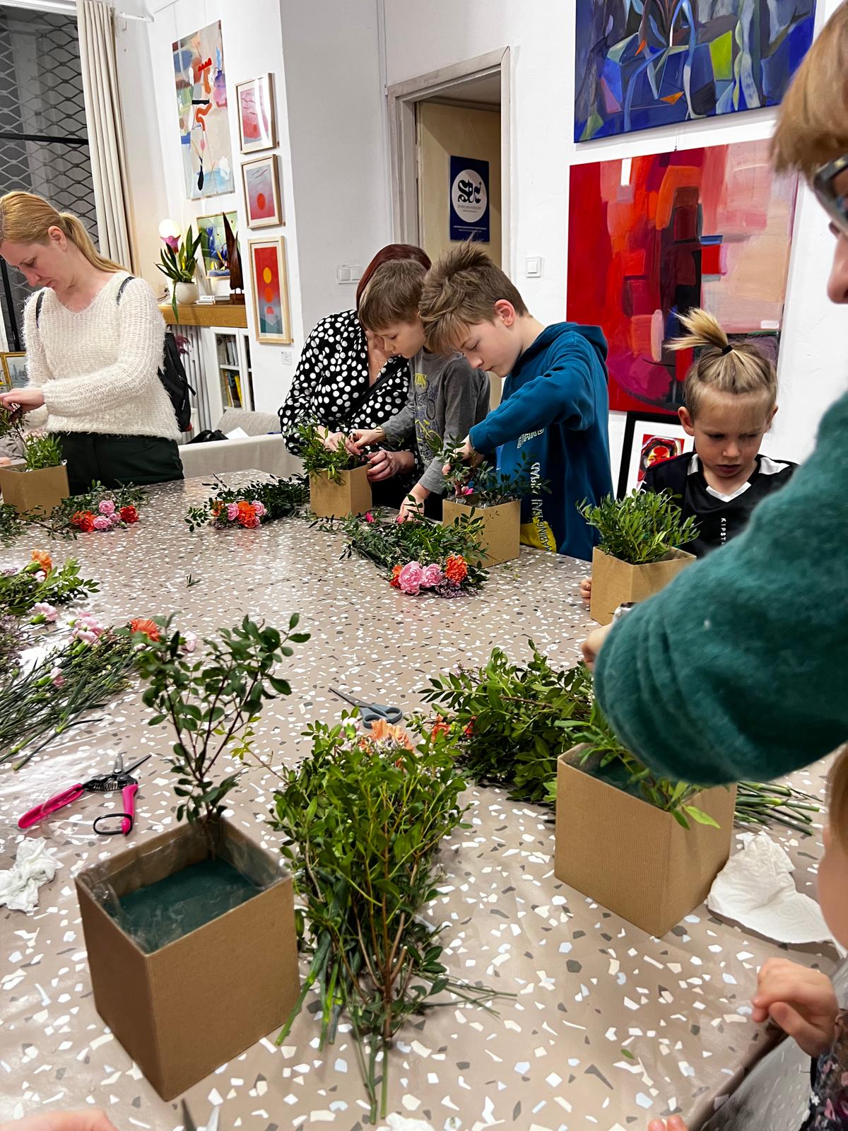 Dzieci i dorośli tworzą kwiatkowe kompozycje w kartonowych pudełkach podczas warsztatu florystycznego w galerii sztuki.