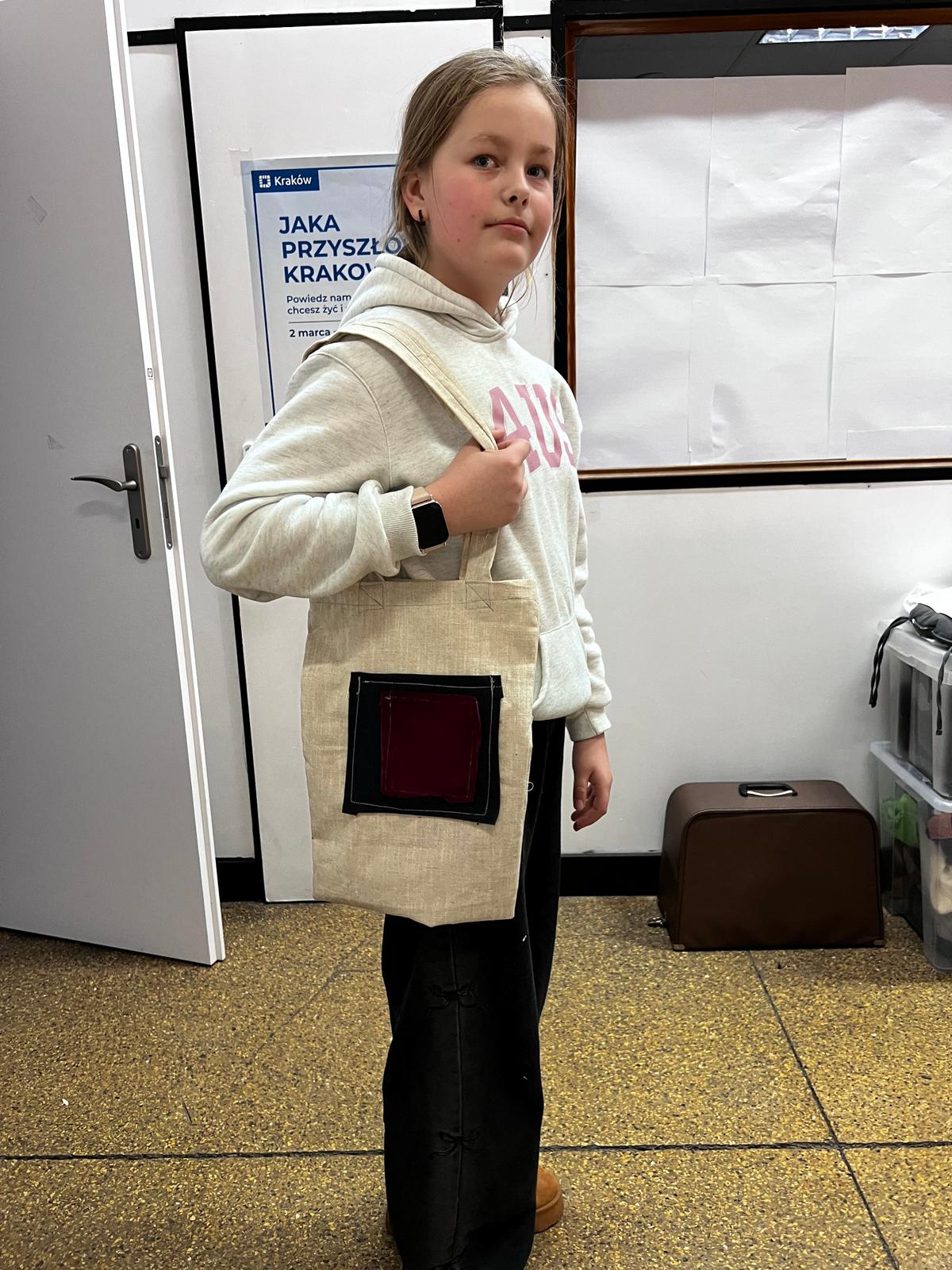 Dziewczynka w beżowej bluzie i kamizelce stoi w holu, a za nią widać plakat reklamowy dotyczący Krakowa.