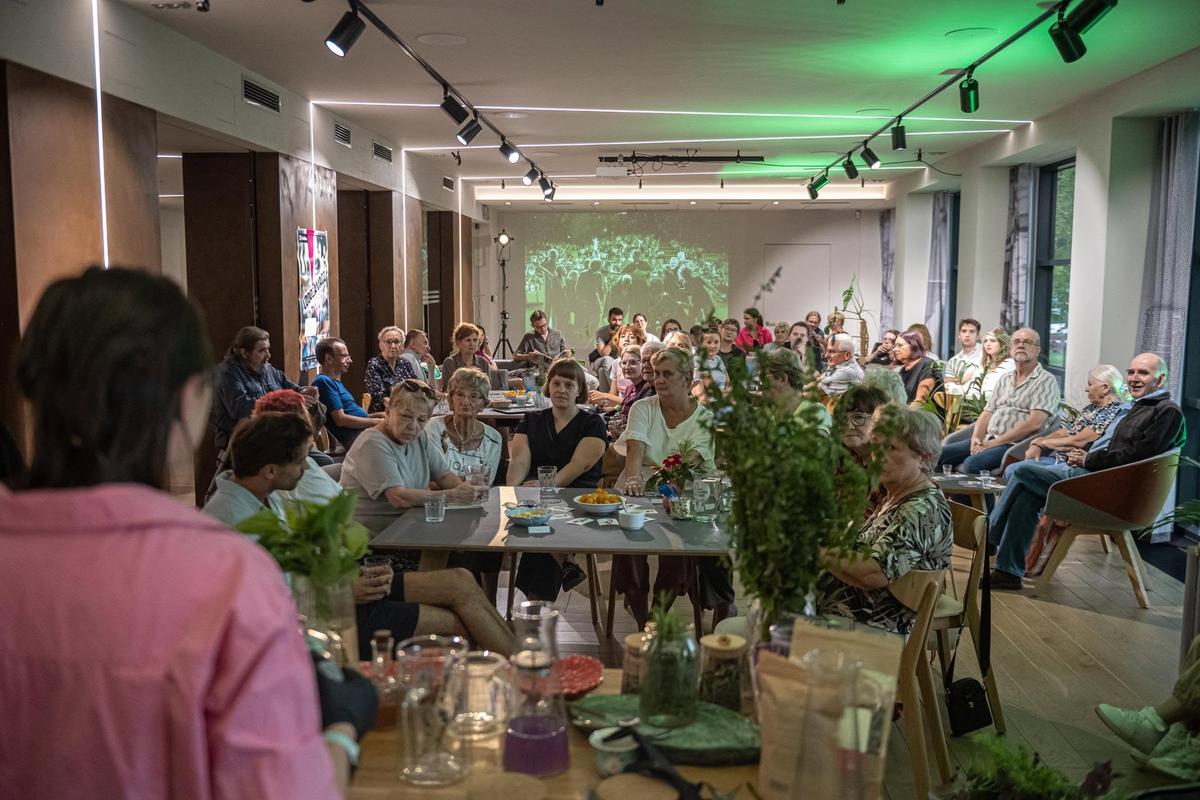 Uczestnicy degustacji naparów sieddzą w nowoczesnej sali, otoczeni zielonymi roślinami, słuchając prelekcji o roślinnych smakach i zapachach.