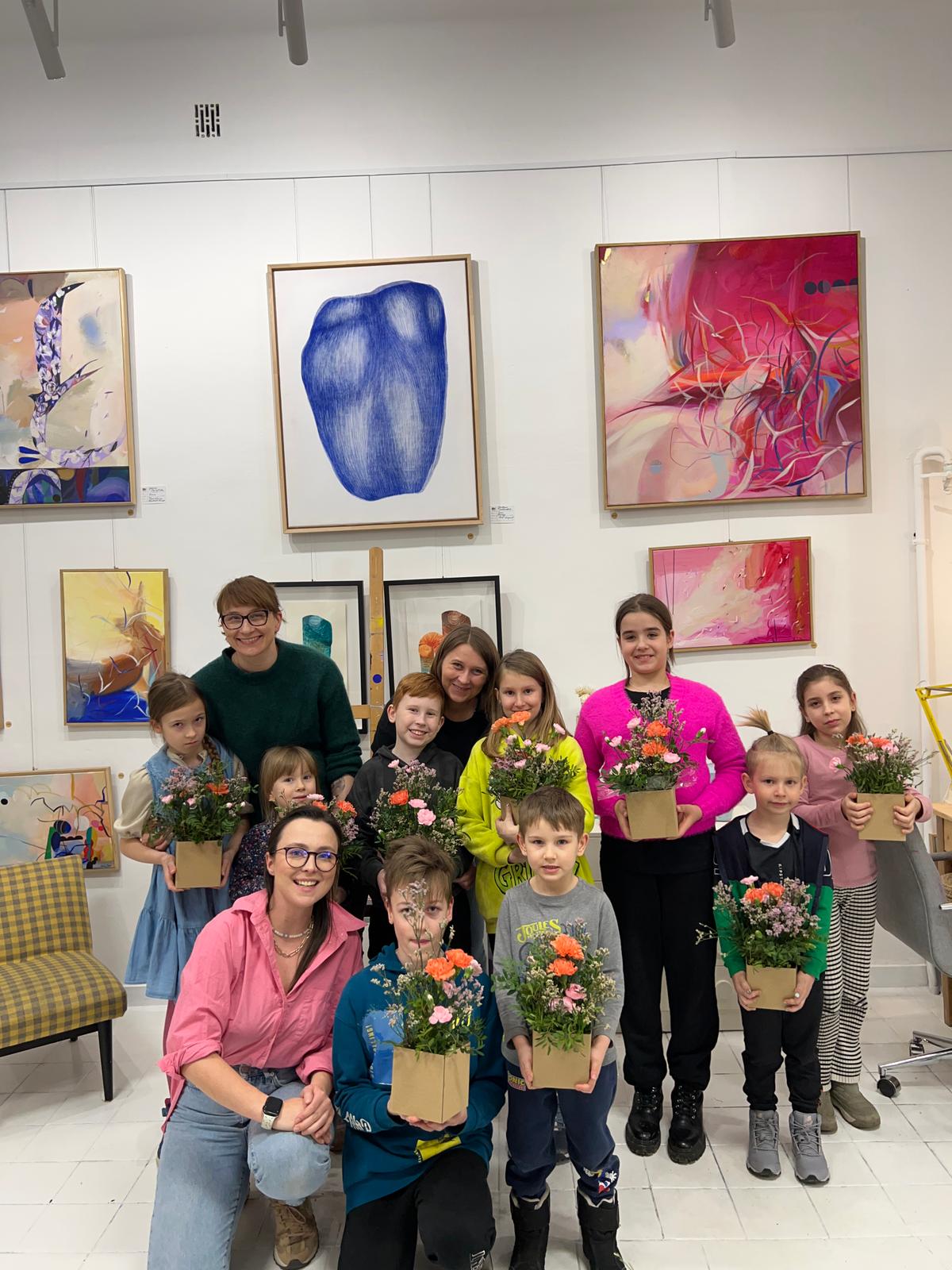 Grupa dzieci i dorosłych pozuje w galerii sztuki, trzymając doniczki z kwiatami przed ścianą z obrazami.