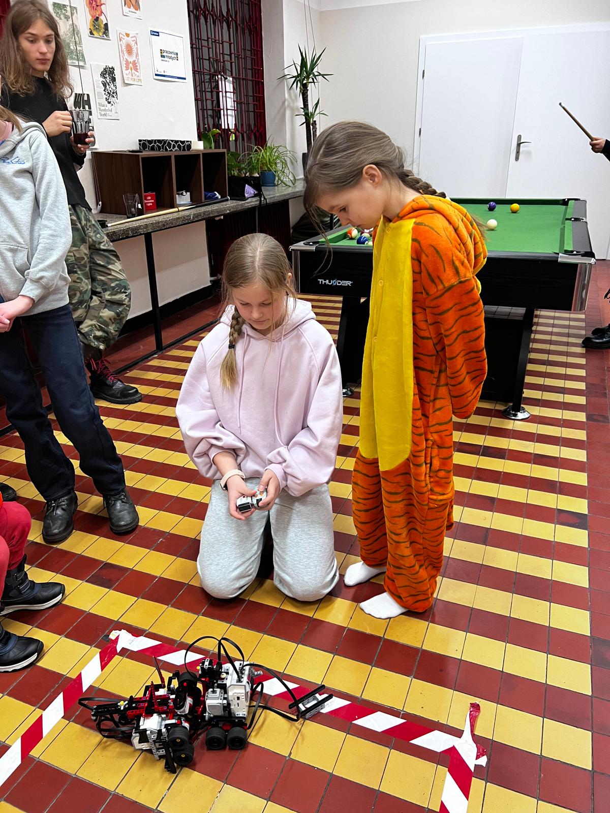 Dziewczyna w różowej bluzie steruje robotem na podłodze, podczas gdy inne dzieci obserwują eksperyment w sali rekreacyjnej.