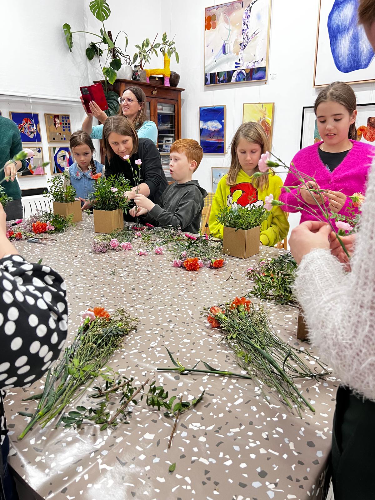Dzieci uczestniczą w warsztacie florystycznym, tworząc kompozycje z kwiatów w drewnianych doniczkach pod opieką instruktorki.