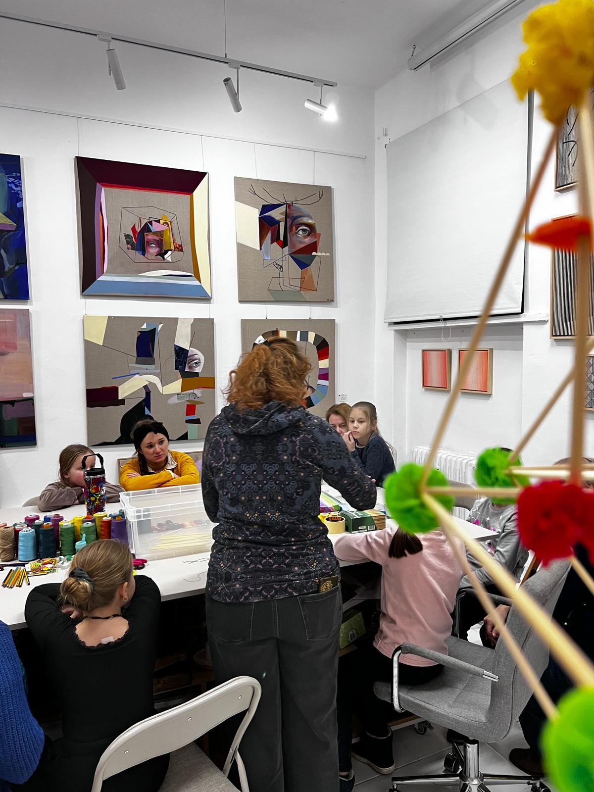 W galerii sztuki instruktorka prowadzi warsztaty twórcze dla grupy dzieci siedzących przy stołach z materiałami plastycznymi.