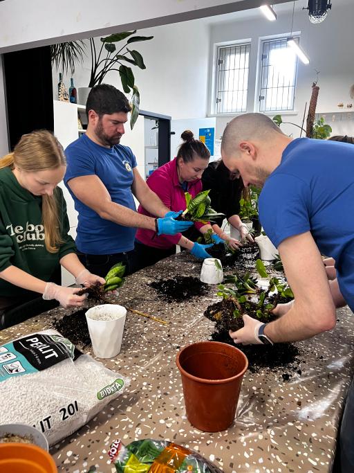 Grupa osób w niebieskich koszulkach sadzą rośliny w ziemi na stole podczas warsztatu edukacyjnego.