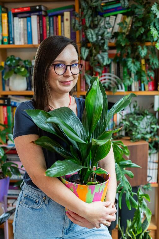 Kobierta w okularach trzyma potted roślinę z zielonymi liśćmi na tle biblioteki pełnej kolorowych książek.