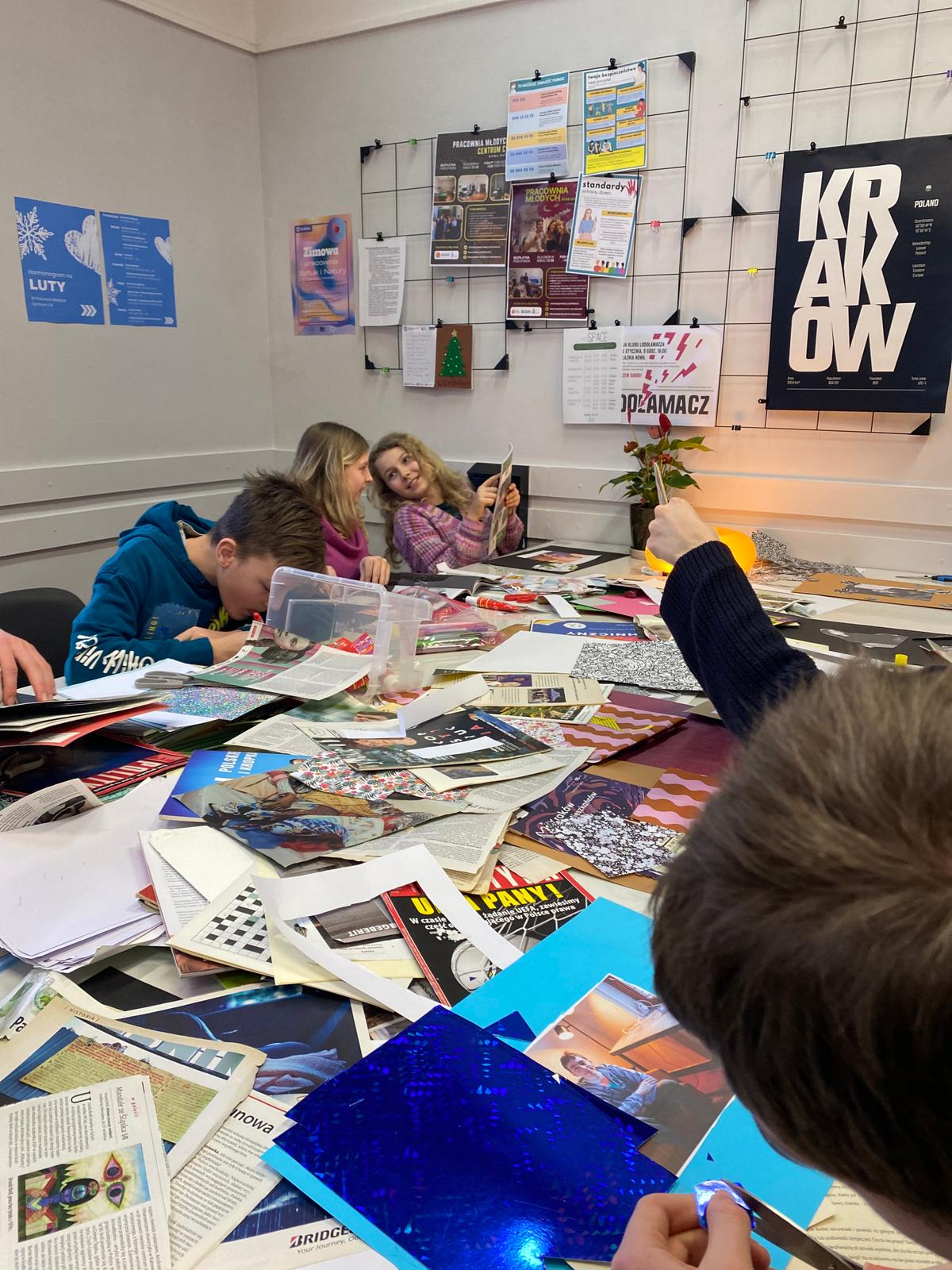 Grupa dzieci siedzi przy stole i pracuje z magazynami i materiałami do collage'u w sali klasowej.