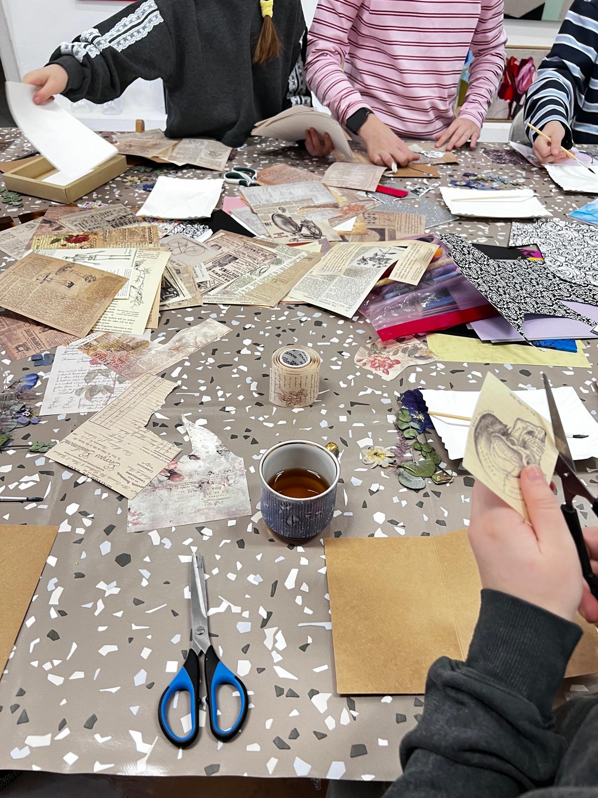 Grupa dzieci pracuje nad kreatywnym projektem z papierem, zdjęciami i narzędziami do cięcia na stole.