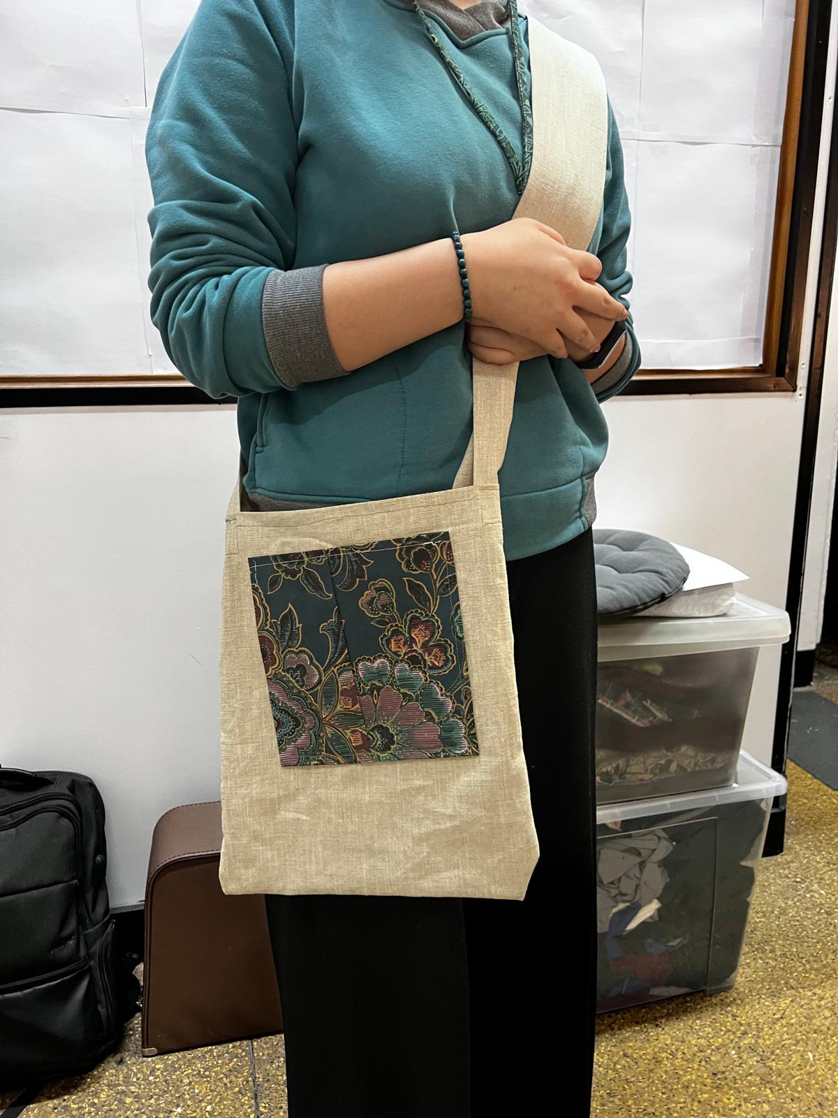 Kobieta w zielonej bluzie prezentuje beżową torbę na ramię z dekoracyjnym kwiatowym panelem.