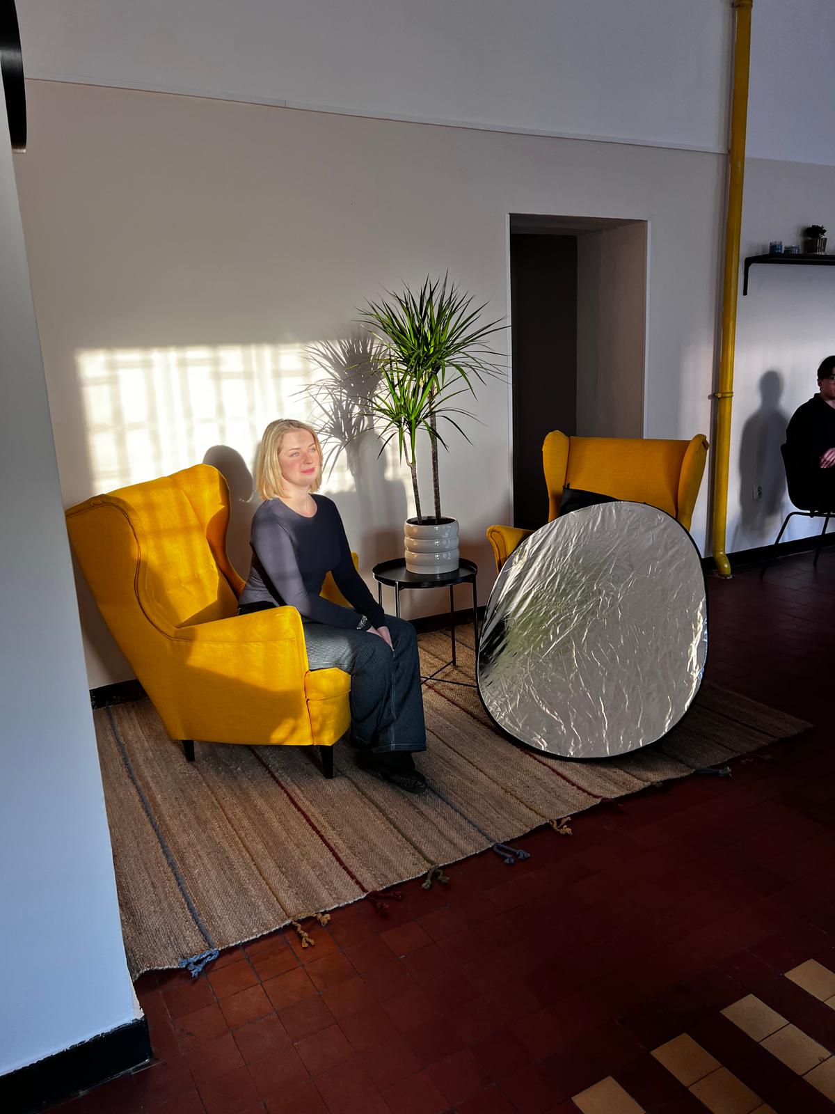 Kobieta siedzi na żółtym fotelu w nowoczesnym wnętrzu z rośliną i reflektorem oświetlającym białą ścianę.