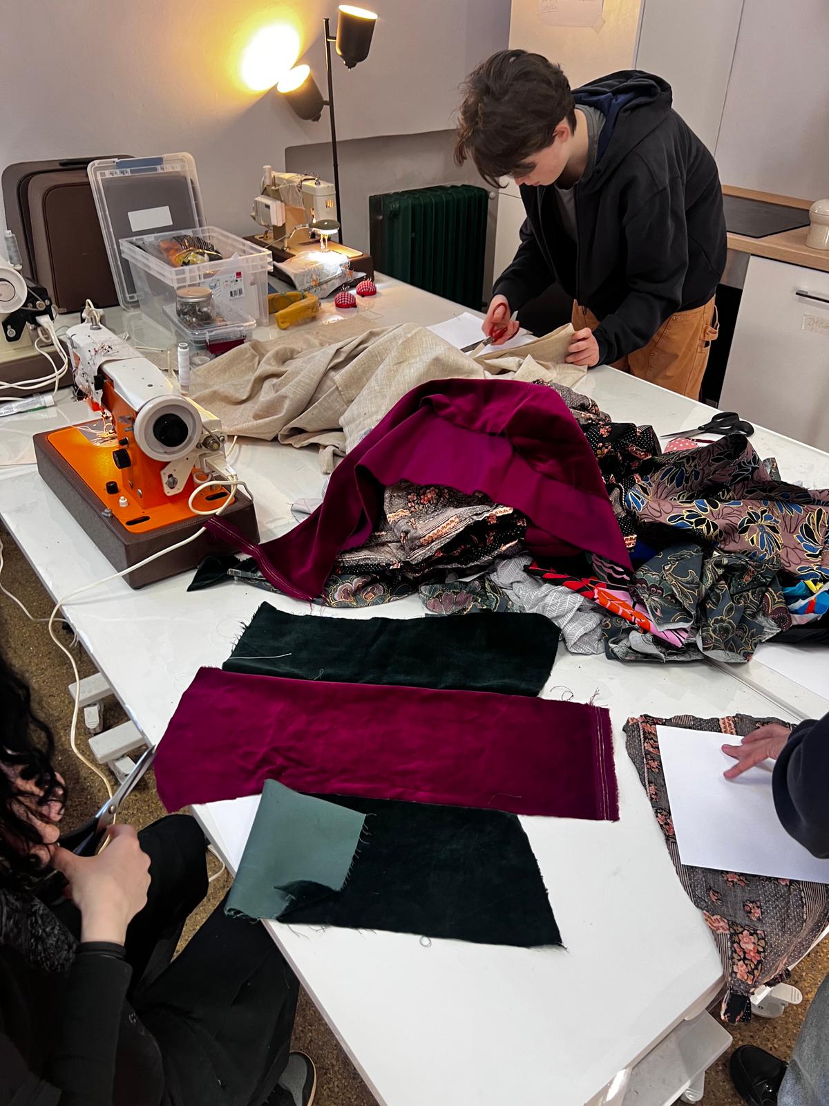 Osoby pracują przy stole z tkaninami i urządzeniami do szycia w warsztacie krawieckim.