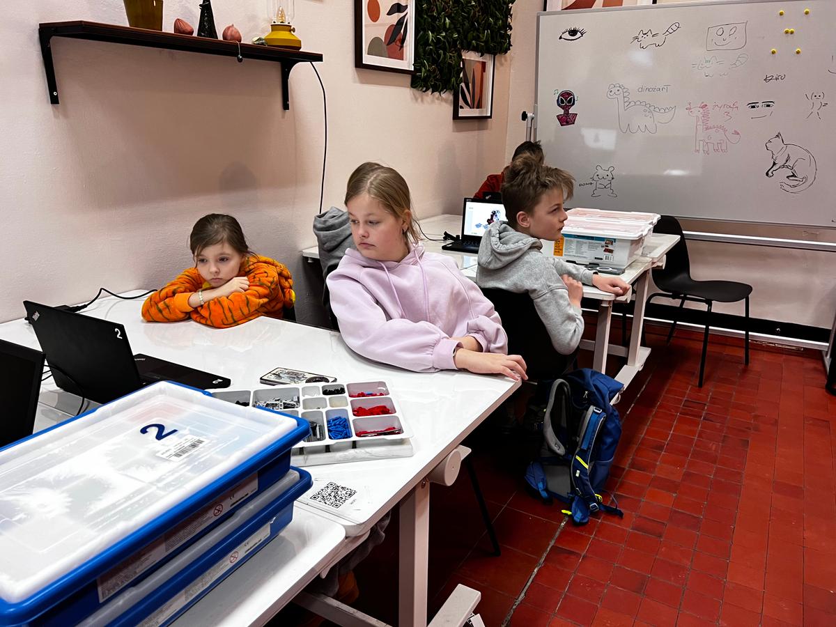 Dzieci siedzą przy stołach w klasie, pracując przy komputerach i materiałach edukacyjnych podczas lekcji.