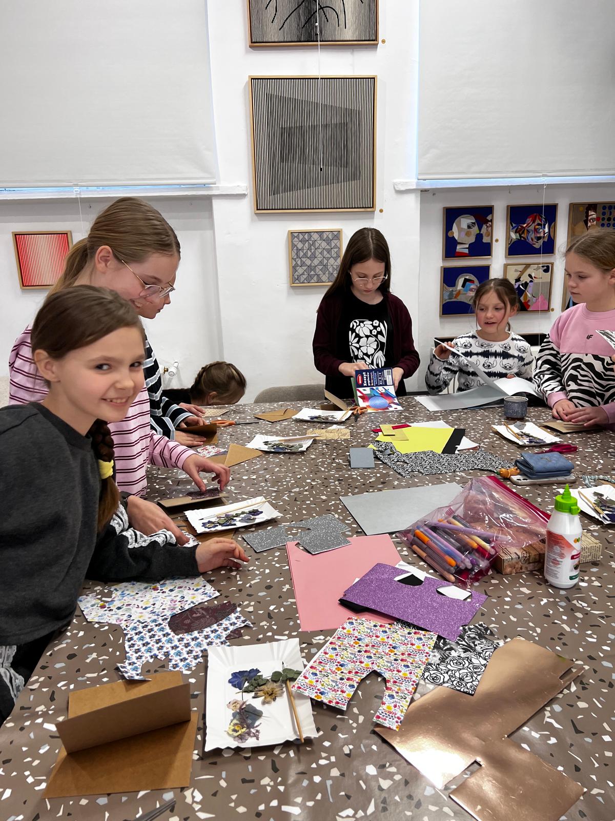 Grupa młodych dziewczyn zajmuje się pracą plastyczną przy stole, otoczona kolorowymi materiałami i papierami do tworzenia.