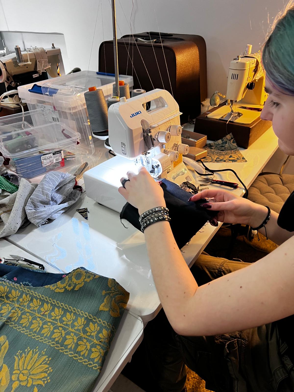 Kobieta szyje na maszynie do szycia w pracowni pełnej materiałów, narzędzi i urządzeń krawieckich.