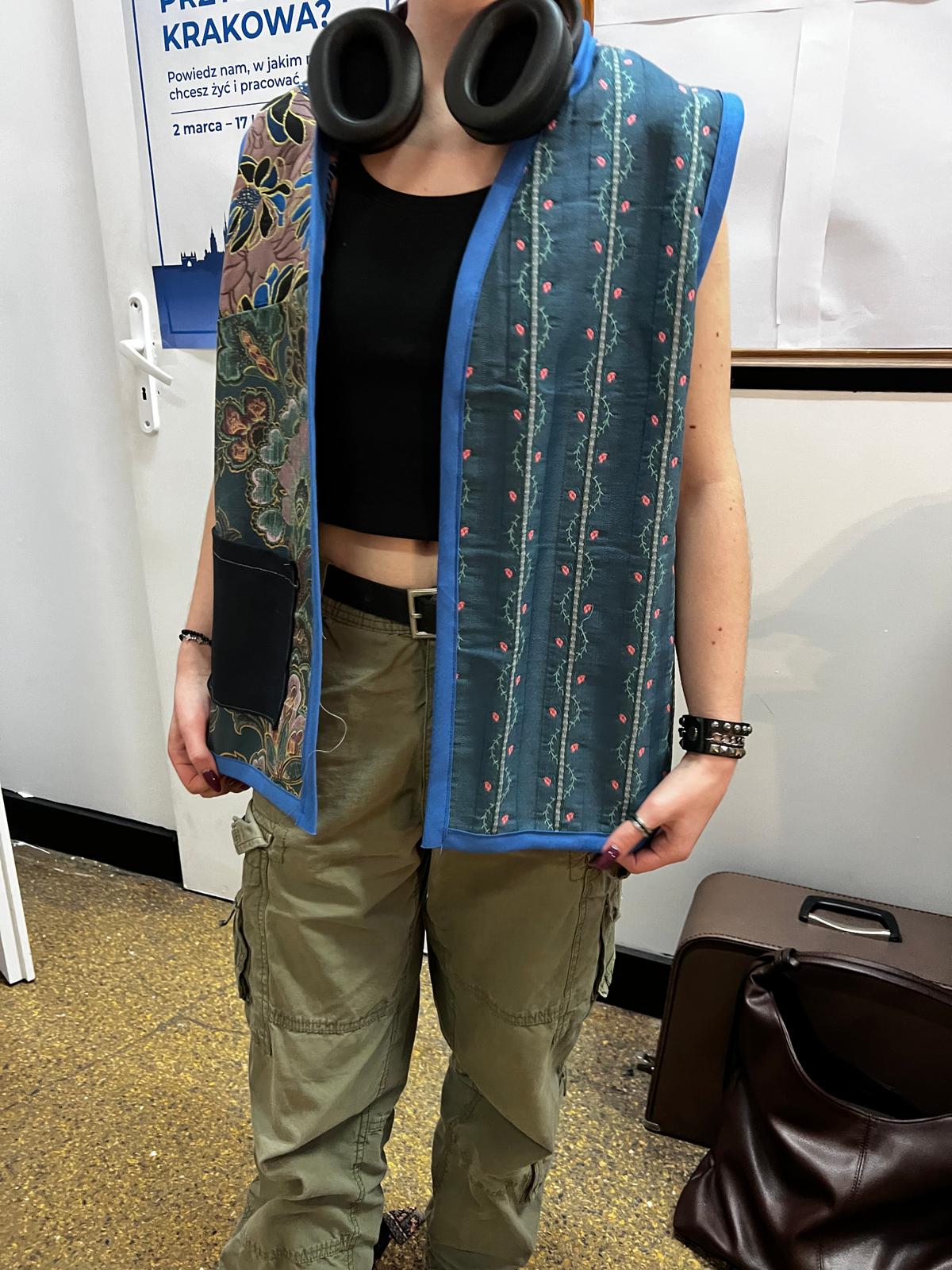 Osoba w czarnym topie, khaki spodniach i wzorzystej kamizelce z niebieskim obramowaniem, ubrana w słuchawki na głowie.