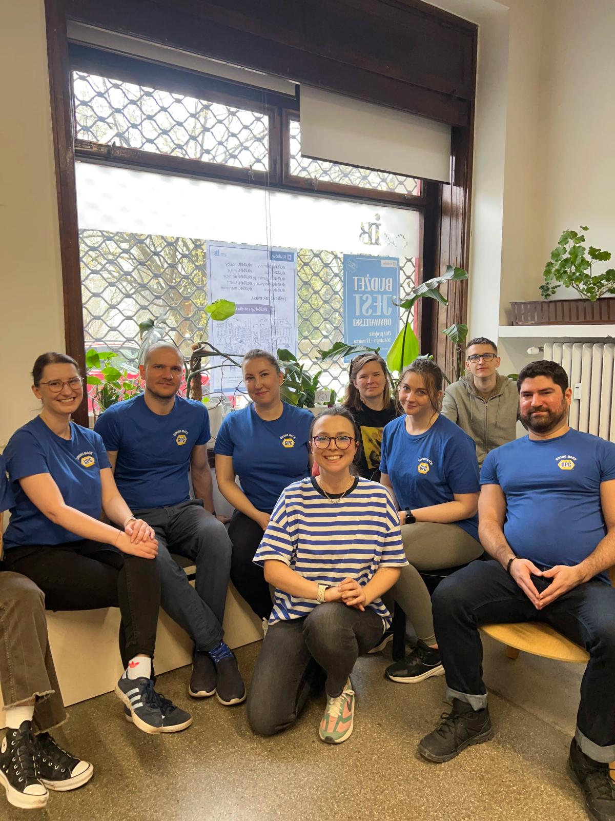 Grupa osób w niebieskich koszulkach pozuje do zdjęcia grupowego w biurze z roślinami i plakatami na tle.Z przodu Prezeska fundacji Czerwony Krakowiak