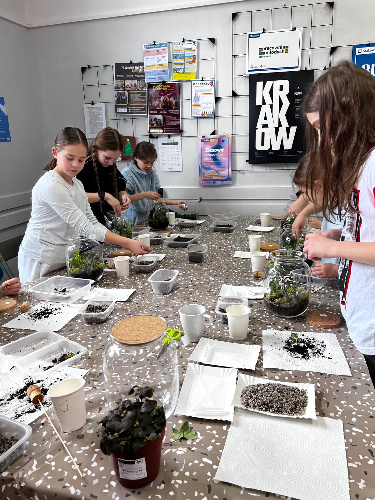 Dzieci pracują przy stole, sadzą rośliny w słoikach jako część warsztatu edukacyjnego w klasie.