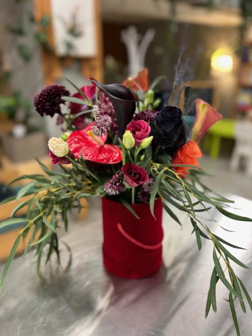 Elegancki bukiet kwiatów w czerwonym wazonie z ciemnymi różami, anturium i zielonymi gałązkami.