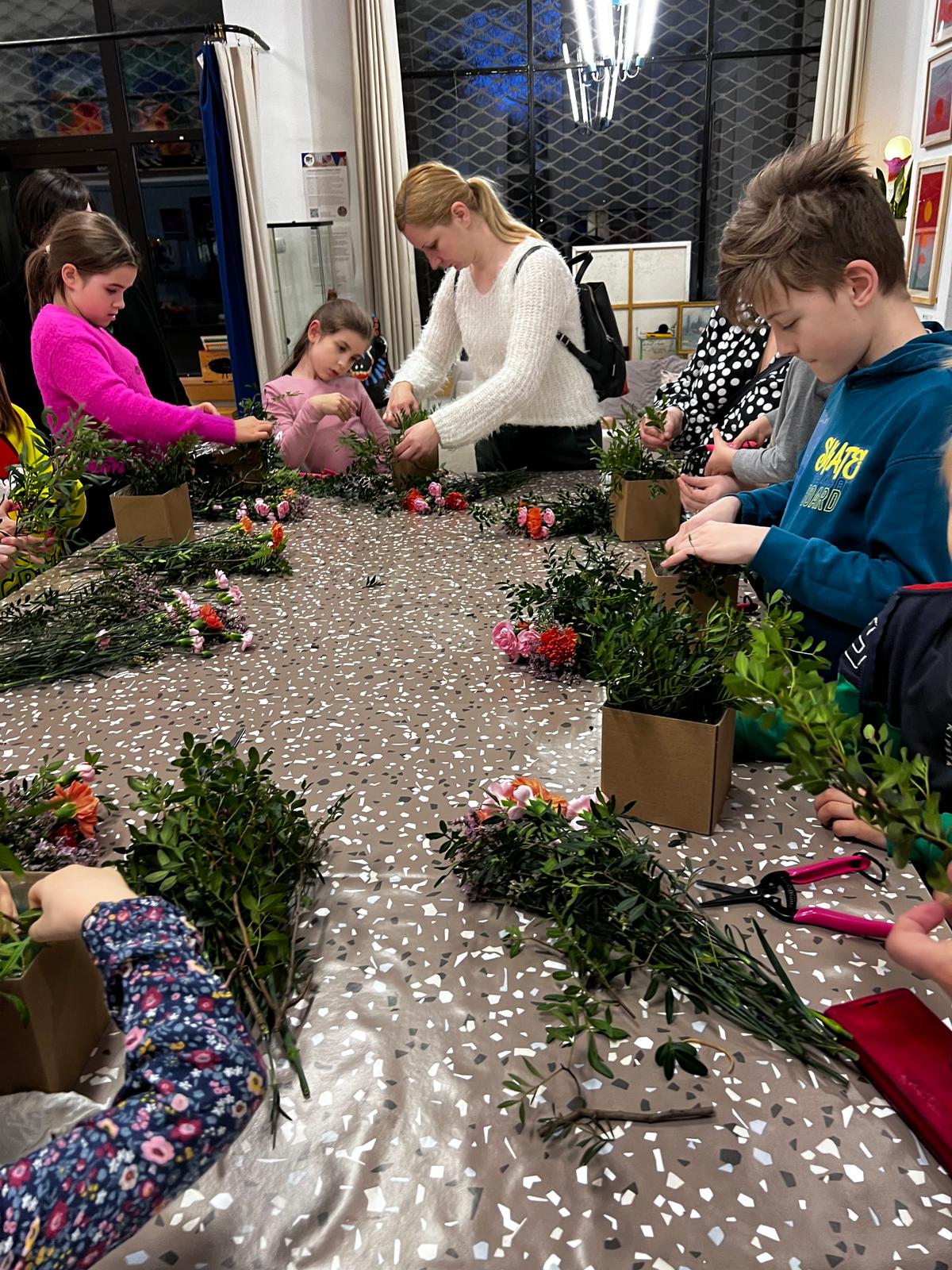 Grupa dzieci uczestniczy w warsztatach florystycznych, przygotowując kompozycje kwiatowe przy pomocy dorosłej instruktorki.