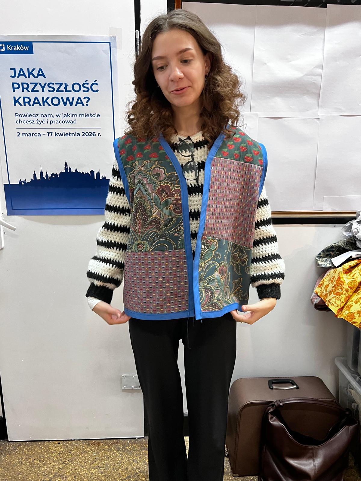 Kobieta prezentuje kolorową kamizelkę z ornamentami na tle plakatu promocyjnego dotyczącego możliwości pracy w Krakowie.