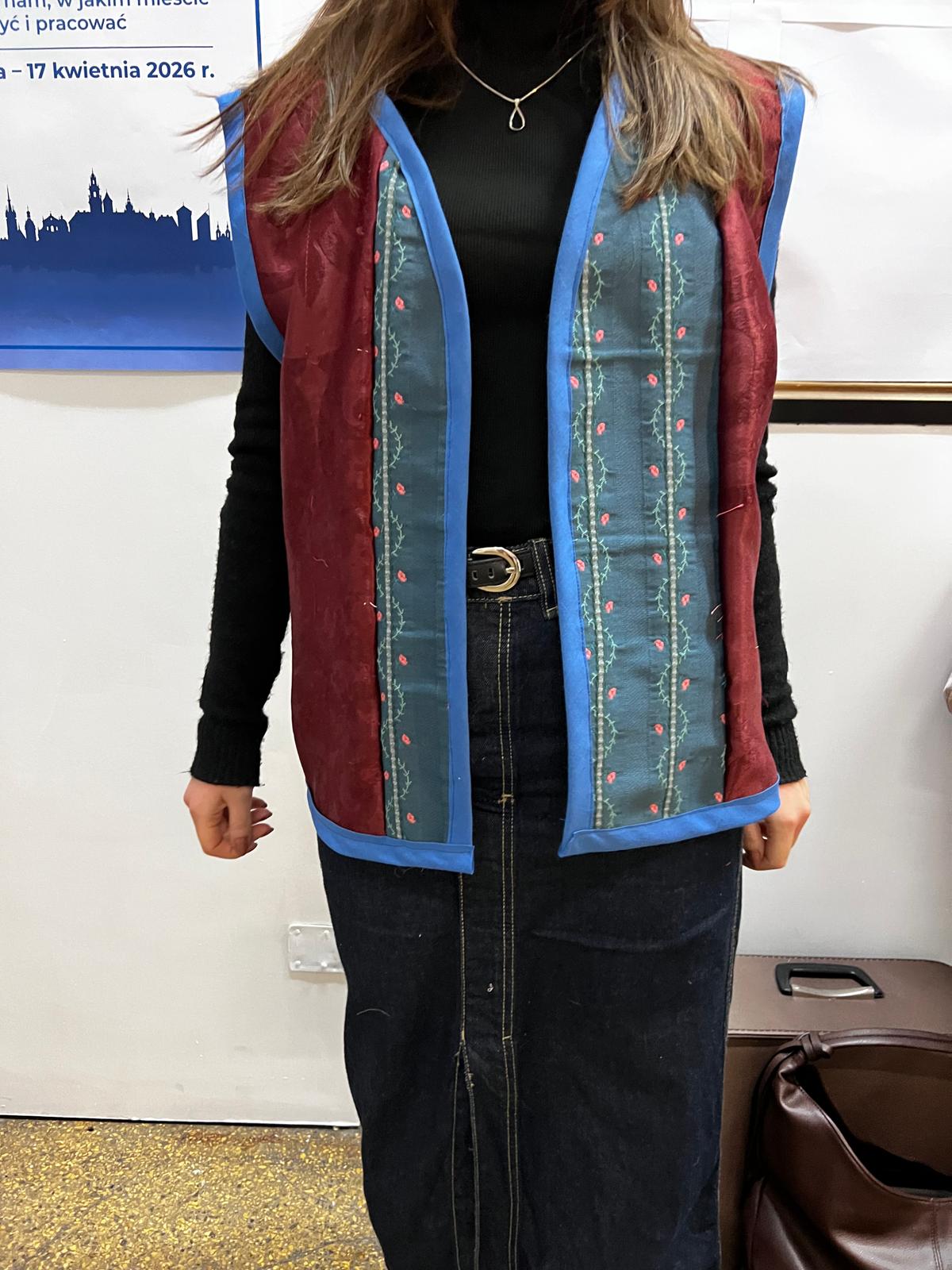 Kobieta w tradycyjnym wielokolorowym bezrękawniku z niebieską lamówką, czarną bluzką i jeansami.