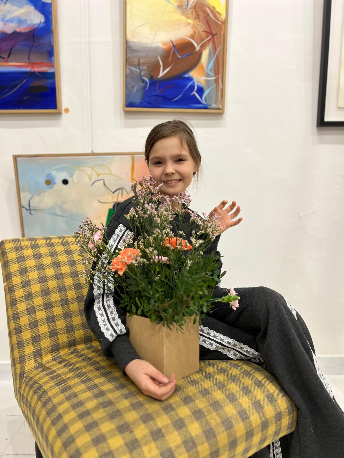 Uśmiechnięta dziewczyna siedzi na żółtym krześle, trzymając bukiet kolorowych kwiatów w kartonowym pojemniku.