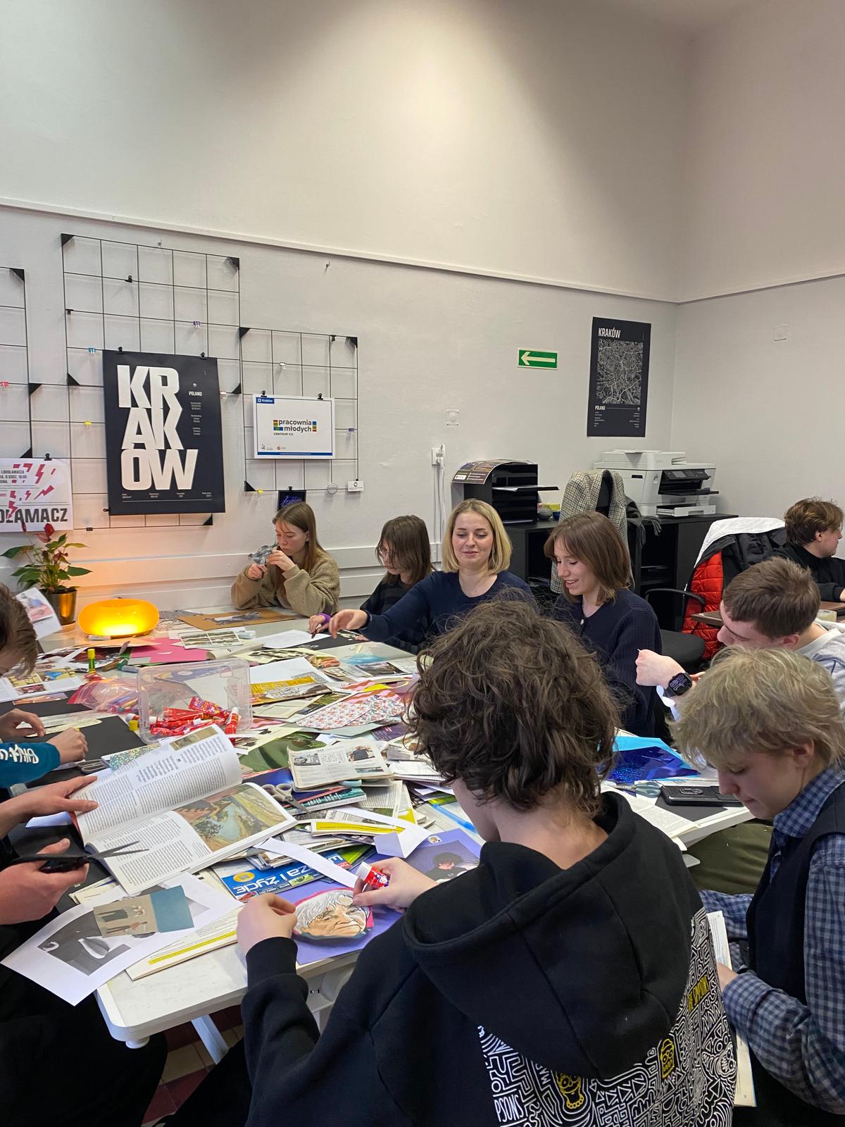 Grupa osób siedzi wokół stołu pełnego magazynów i materiałów w biurze z plakatem Krakowa na ścianie.
