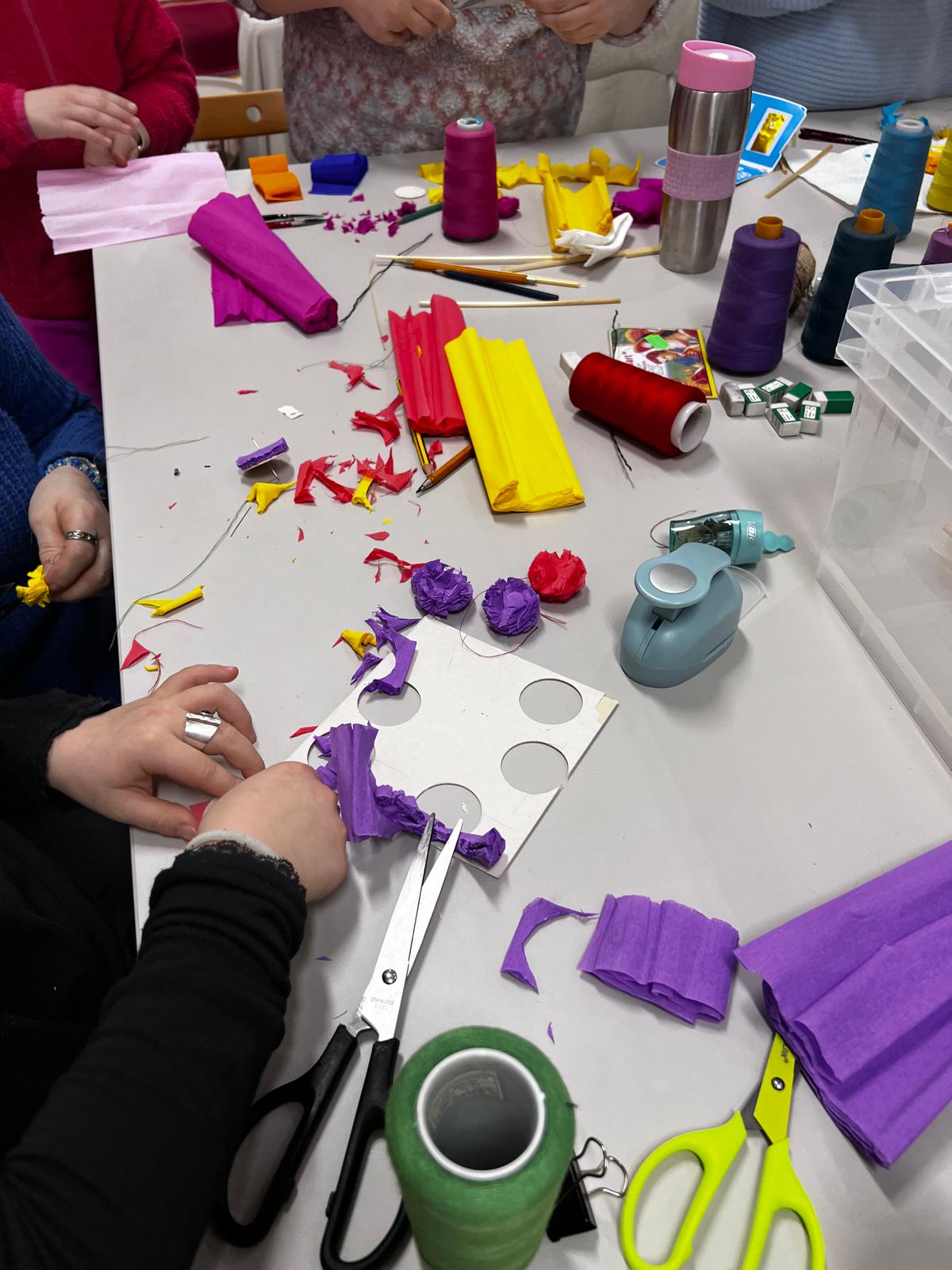 Grupa dzieci wykonuje kolorowe papierowe dekoracje, używając nożyczek, taśmy i kolorowych tkanek na białym stole roboczym.