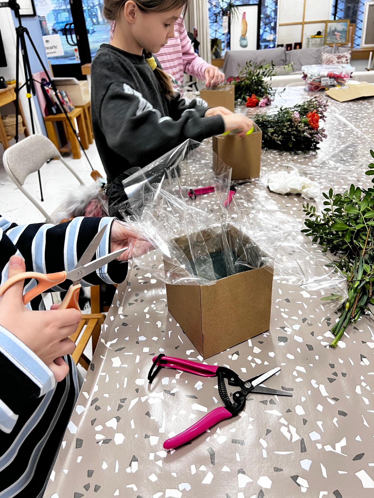 Dzieci przygotowują kwiaty do bukietów, owijając je folią i przycinając łodygi nożyczkami w pracowni florystycznej.