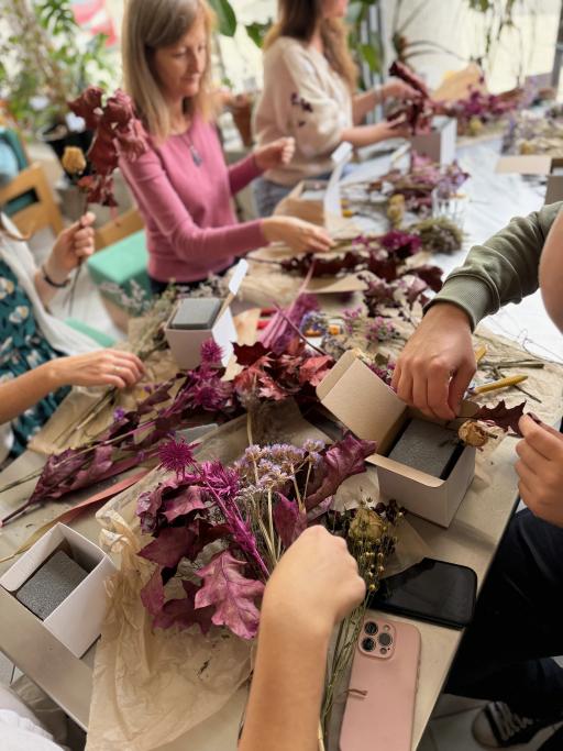 Grupa osób tworzy bukiety z wysuszonych kwiatów w odcieniach burgundu i fioletu przy drewnianym stole.