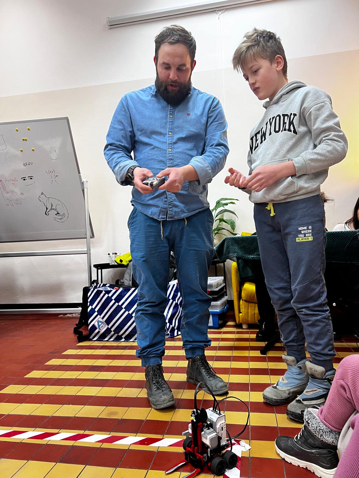 Mężczyzna w niebieskiej koszuli i chłopiec w szarej bluzie demonstrują małego robota na kolorowej podłodze w klasie.