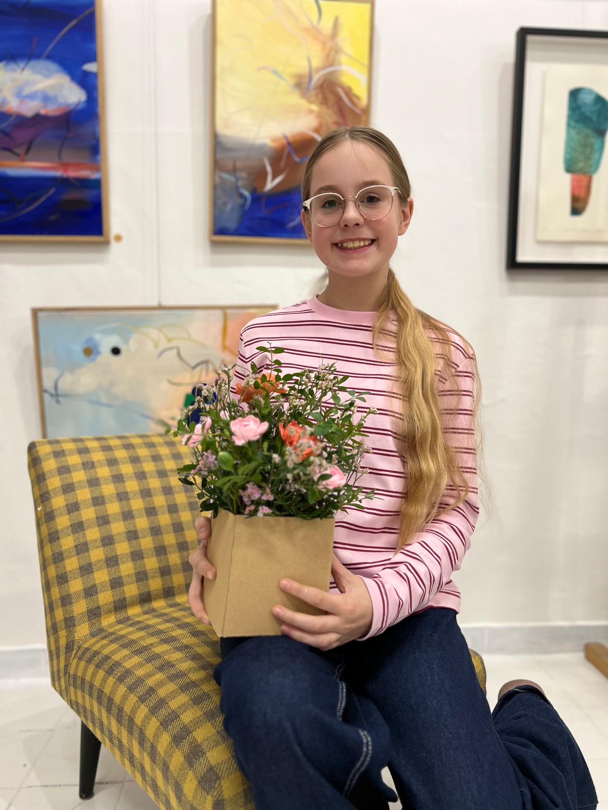 Uśmiechnięta dziewczyna w okularach siedzi na żółto-niebieskim krześle, trzymając doniczkę z kolorowymi kwiatami.
