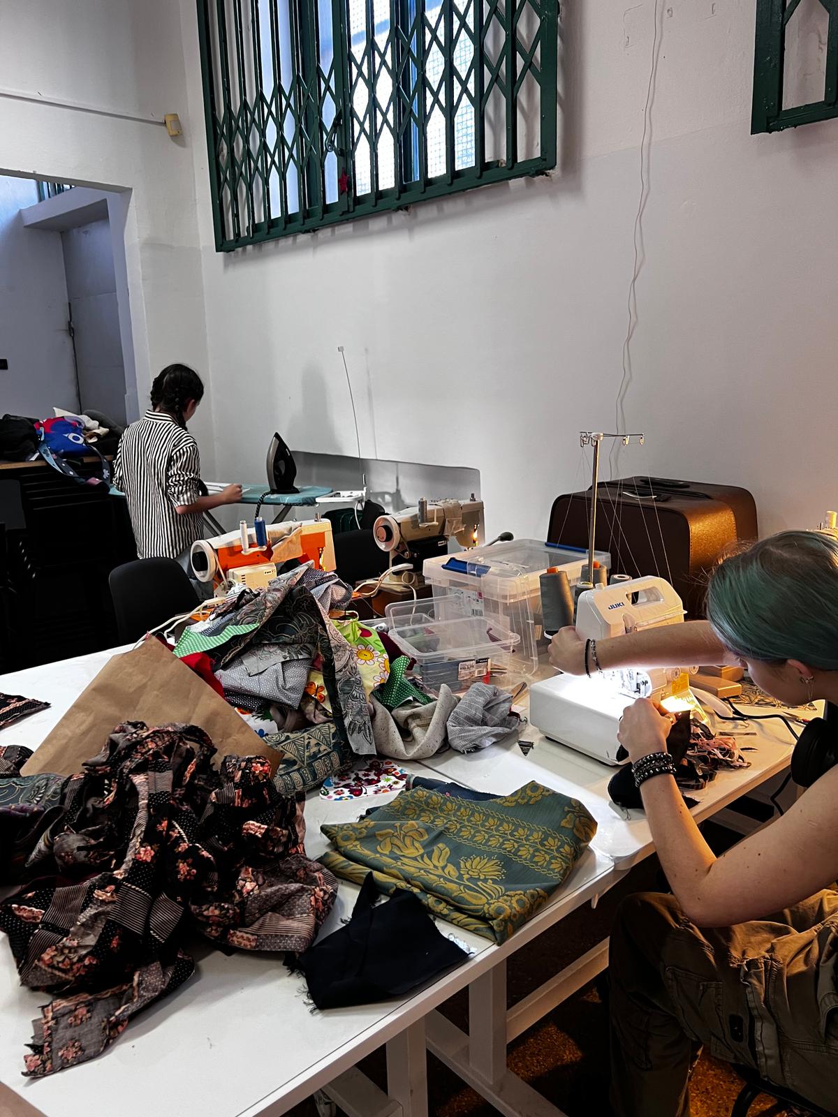 Dwie osoby pracują przy stołach pełnych tkanin, nici i narzędzi do szycia w pracowni rzemiosła.