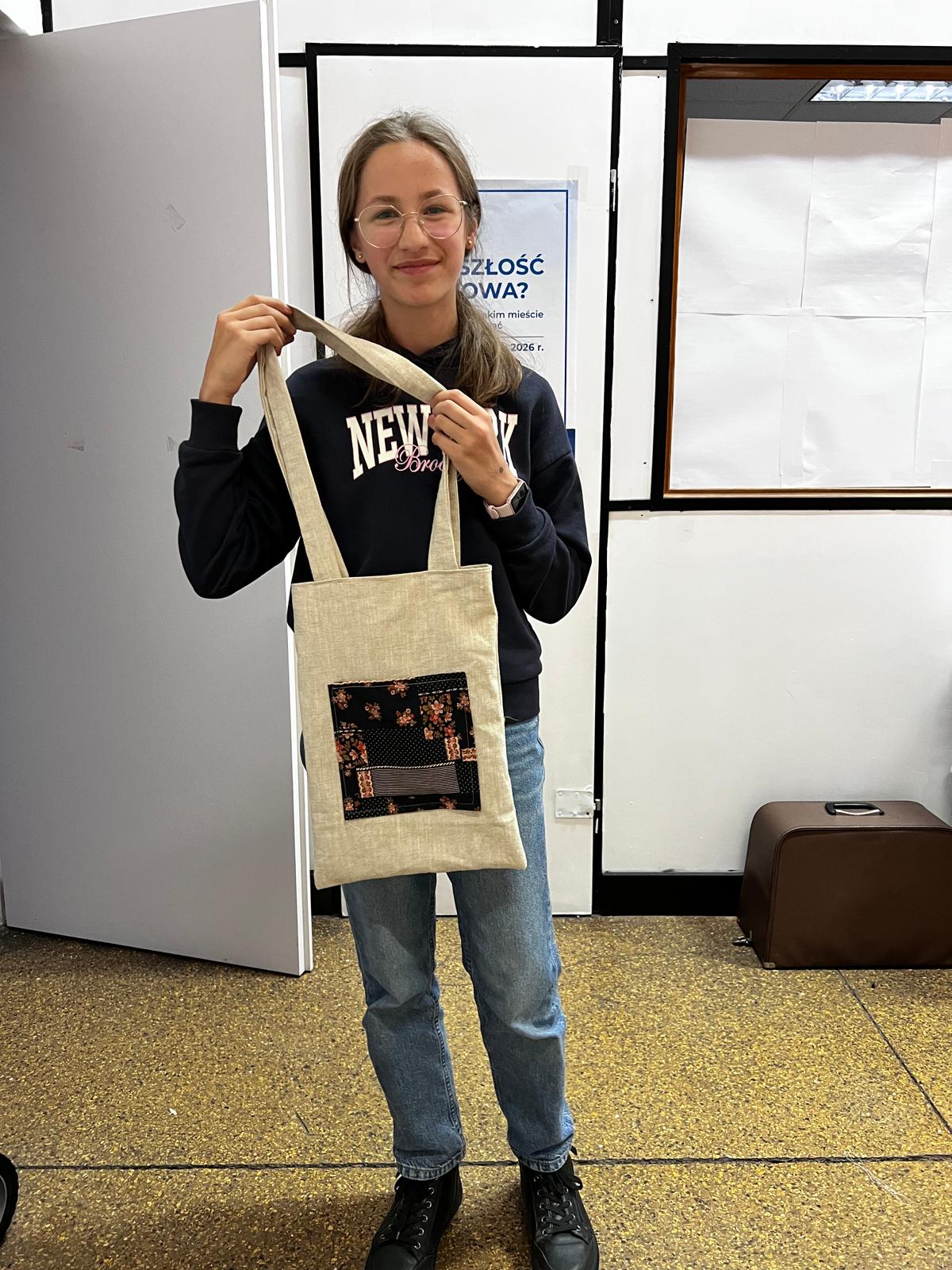 Dziewczyna w okularach i czarnej bluzie prezentuje beżową torbę na ramię z kolorową aplikacją.