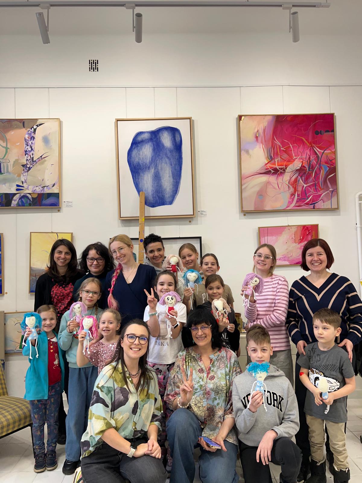 Grupa dzieci i dorosłych pozuje do zdjęcia w galerii sztuki, trzymając wykonane przez siebie lalki.