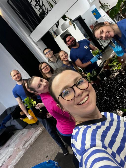 Grupa uśmiechniętych ludzi pozuje do selfie w pomieszczeniu z roślinami, ubrani głównie w niebieskie ubrania.