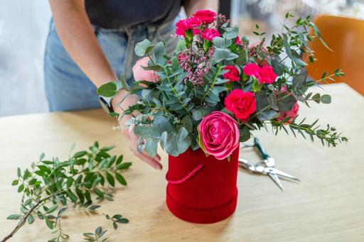 Osoba w niebieskich dżinsach przygotowuje bukiet różowych róż w czerwonym pudełku na drewnianym stole.