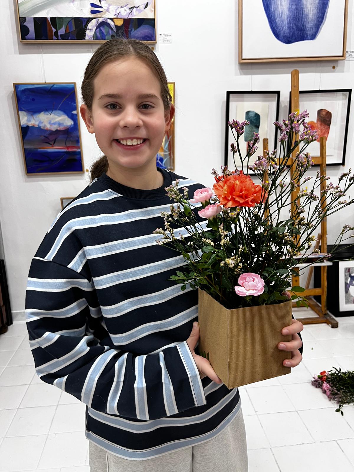 Uśmiechnięta dziewczyna w pasiastym swetrze trzyma wazon z kolorowymi kwiatami w galerii sztuki.