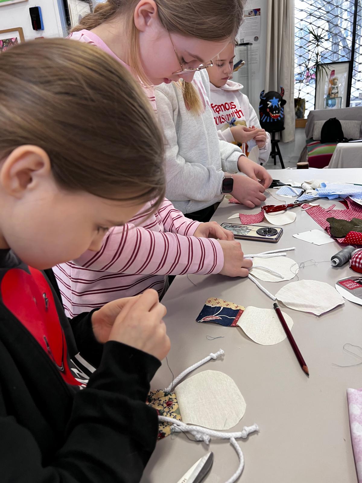 Dzieci wykonują pracę plastyczną przy stole, dekorując materiałowe elementy za pomocą farb i pędzli.