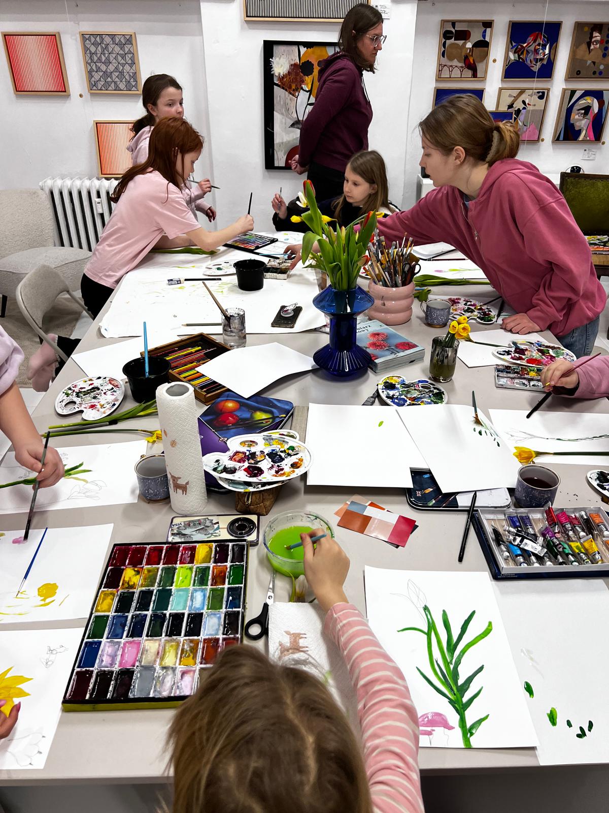 Grupa dzieci i młodzieży uczestniczy w warsztacie malarskim w galerii sztuki, malując akwarelami przy stołach.
