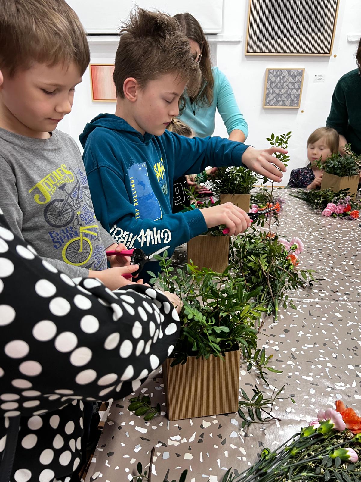 Dzieci uczestniczą w warsztacie florystycznym, przygotowując dekoracyjne kompozycje z kwiatów i roślin w papierowych doniczkach.