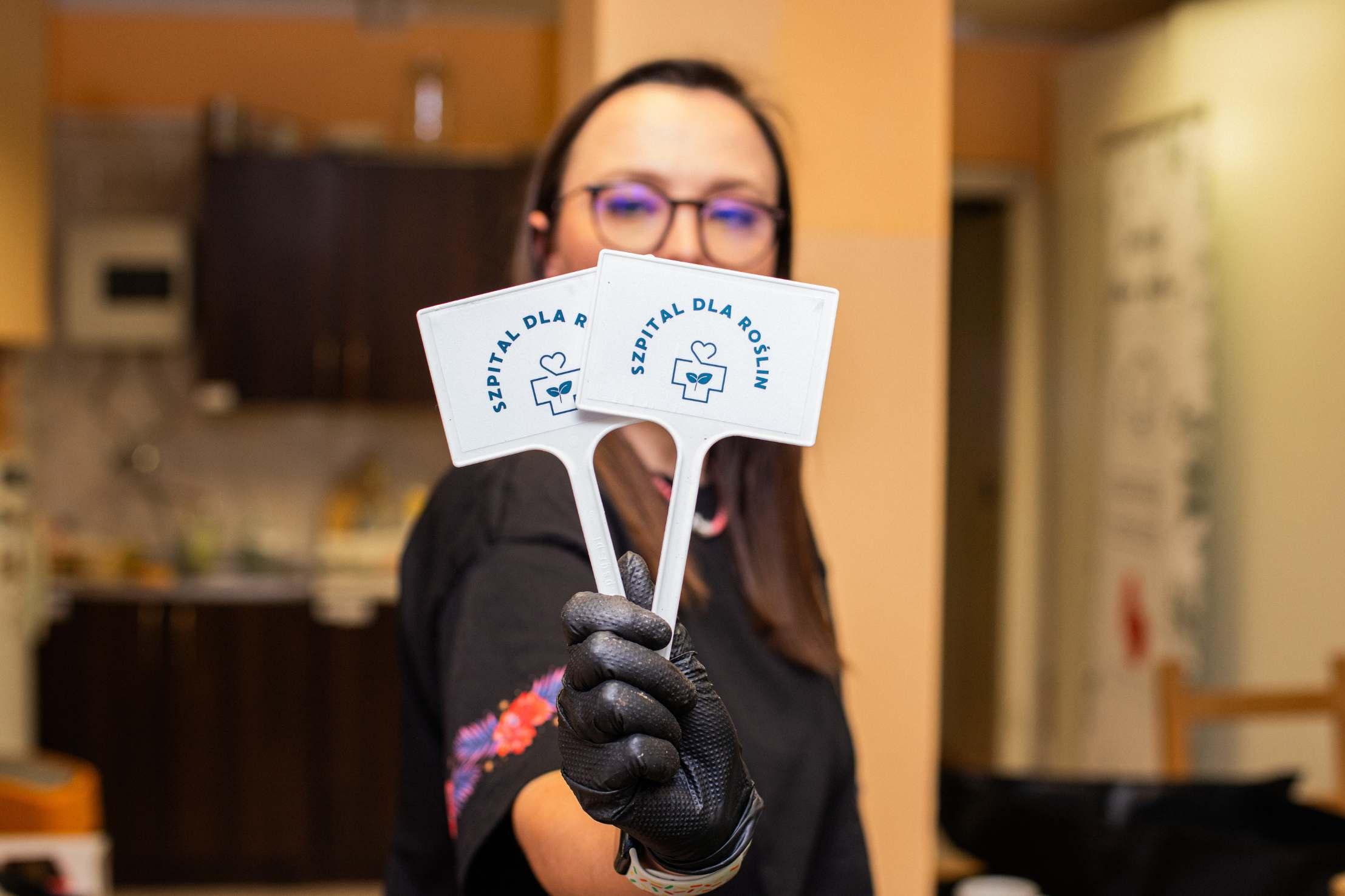 Osoba w okularach i czarnych rękawiczkach trzyma dwie karty z napisem "Szpital dla Röschm" z logiem medycznym.