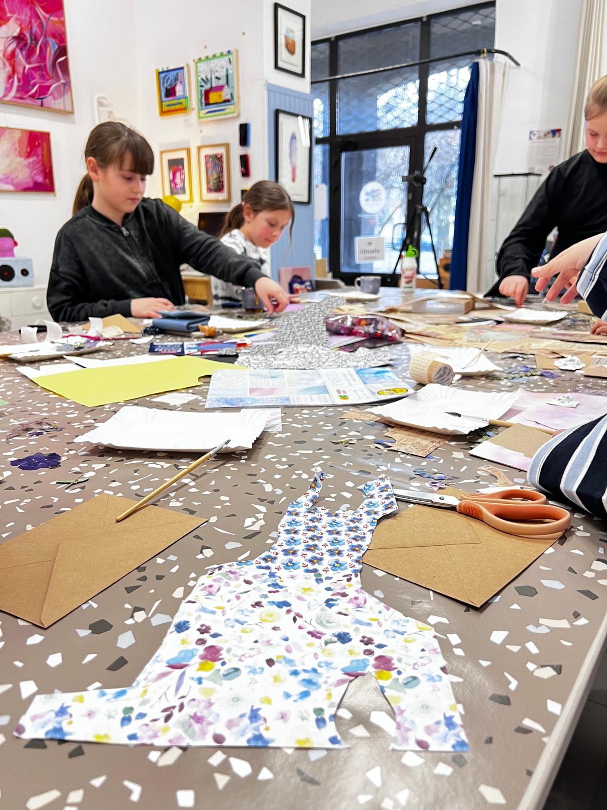 Dzieci uczestniczą w warsztatach artystycznych, tworząc kolorowe projekty z papieru i tkanin przy dużym stole.