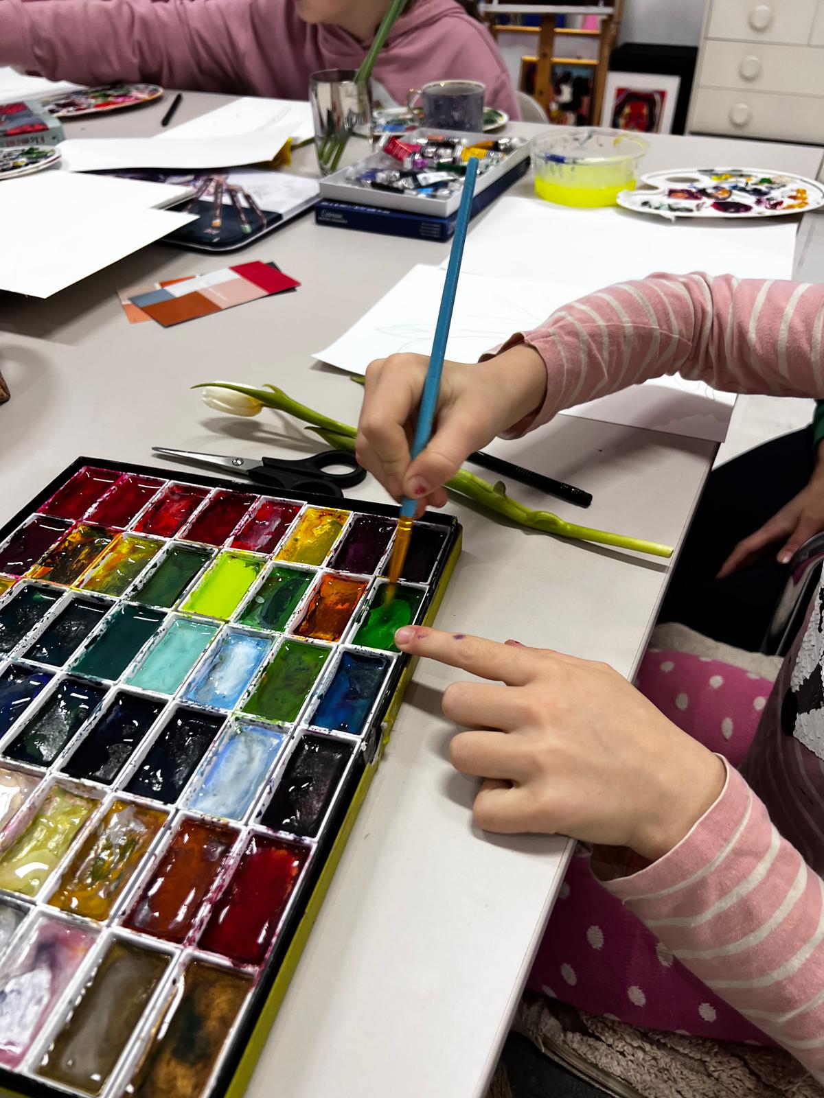 Dziecko maluje pędzlem na papierze, wybierając kolory z rozłożonej palety akwarelowych farb.