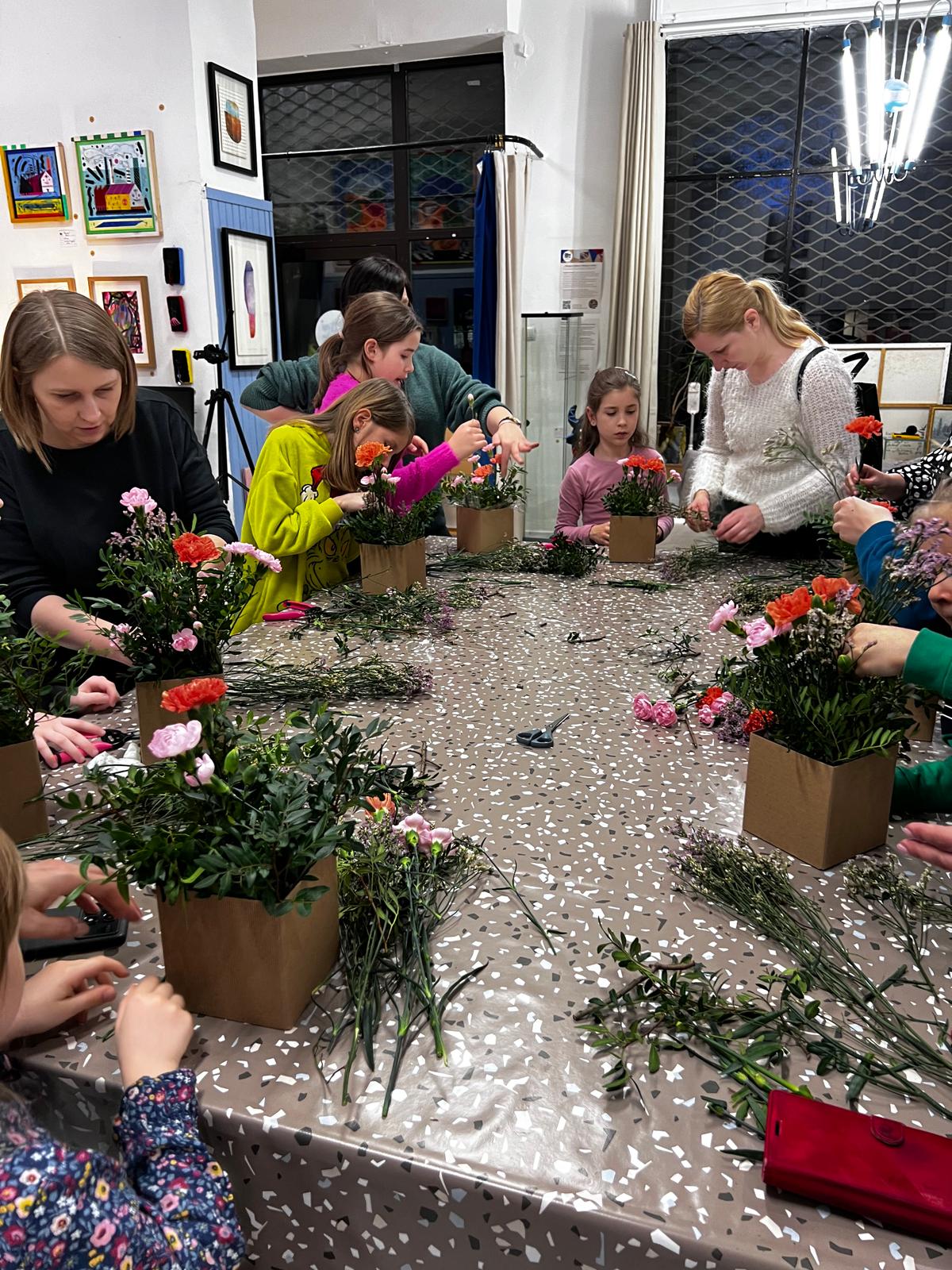 Grupa dzieci i dorosłych uczestniczy w warsztacie florystycznym, arranżując kolorowe kwiaty w doniczkach.